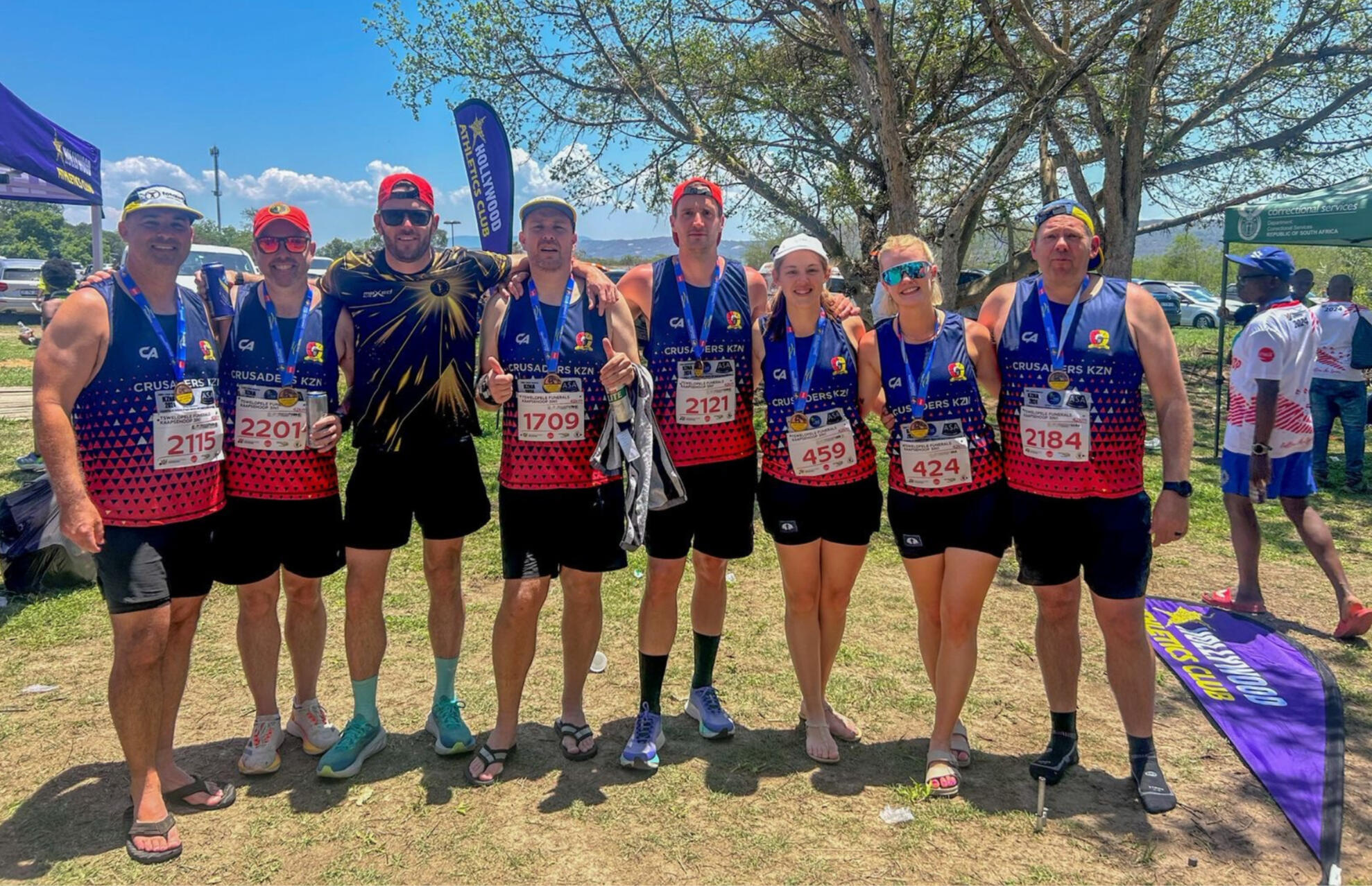 Crusaders Running Club Race Day Crew Group photo of Crusaders Running Club members wearing red and blue race vests, smiling with medals after completing a race on a sunny day at the finish area.